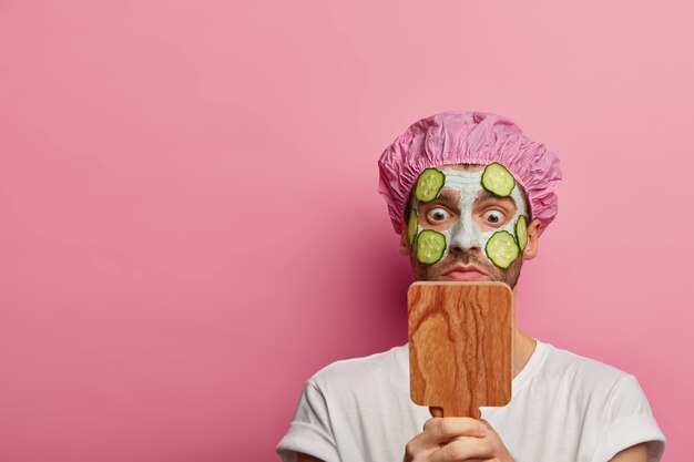 Estupefacto modelo masculino europeo mira al espejo, aplica mascarilla de arcilla con pepinos, tiene tratamientos de belleza en el salón de spa, vestido con camiseta blanca y gorro de baño