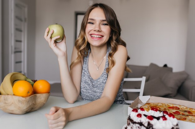 Eufórica señorita divirtiéndose durante el desayuno con manzanas verdes y naranjas. Foto interior de niña caucásica positiva comiendo frutas y pasteles.