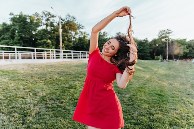 Eufórica señorita en traje de verano jugando con su cabello durante la sesión de fotos en el parque. Tiro al aire libre de linda chica en vestido rojo divirtiéndose en fin de semana.