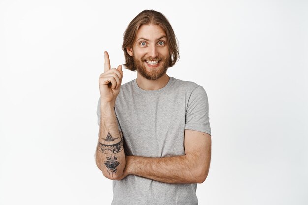 Eureka. Chico rubio guapo sonriente señalando con el dedo hacia arriba, se ve feliz, solución de sugerencia, compartiendo pensamientos, parado en una camiseta gris sobre fondo blanco