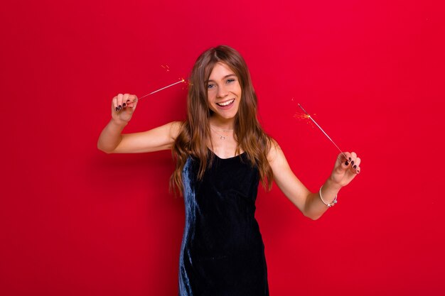 Evento de fiesta brillante de alegre joven feliz en elegante vestido negro celebrando y divirtiéndose sobre fondo rojo aislado con bengalas.