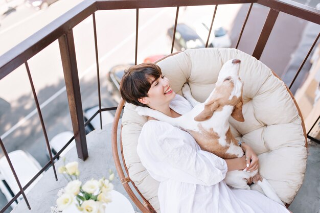 Excelente chica con una sonrisa encantadora disfruta el sábado por la mañana en el balcón con un gracioso perro beagle.