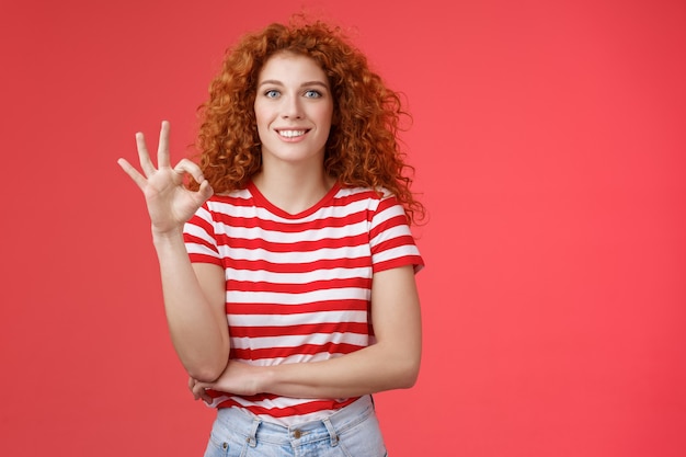 Excelente elección buena. Atractiva mujer rizada pelirroja europea alegre mostrar gesto de aprobación ok ok sonriendo con dientes encantado satisfecho increíble servicio perfecto recomendar producto fondo rojo.