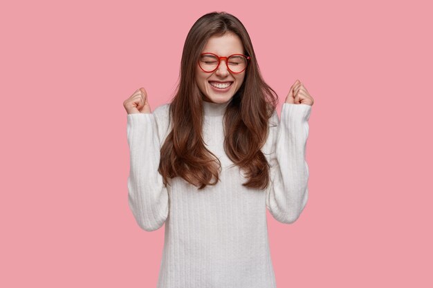 Excesiva mujer joven feliz aprieta los puños, feliz de ganar y ser campeón, lleva gafas ópticas y suéter blanco