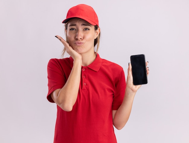 Foto gratuita excitada joven bonita mujer de entrega en uniforme pone la mano en la barbilla y sostiene el teléfono aislado en la pared blanca