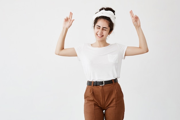 La excitada mujer de cabello oscuro señala el espacio de la copia, cierra los ojos, sonríe ampliamente, publicita. Feliz mujer en camiseta blanca posa contra la pared blanca con área de copia para texto promocional