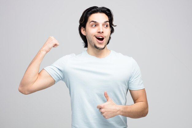 excitado joven apuesto mirando al costado mostrando el pulgar hacia arriba y un gesto fuerte aislado en fondo blanco