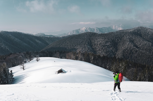 Excursionista macho tomando fotos de una montaña alpina de invierno y una cabaña cubierta de nieve debajo