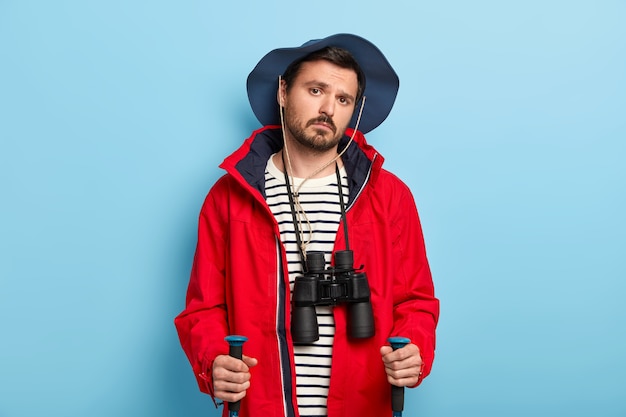 El excursionista masculino joven serio tiene un viaje en el bosque, usa palos de trekking, disfruta del estilo de vida de viaje, usa binoculares en el cuello, usa sombrero casual y chaqueta roja, aislado en la pared azul