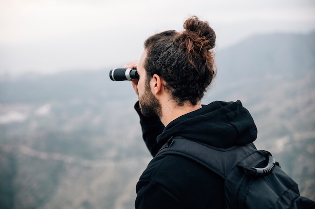 Un excursionista masculino con su mochila mirando a la montaña a través de binoculares