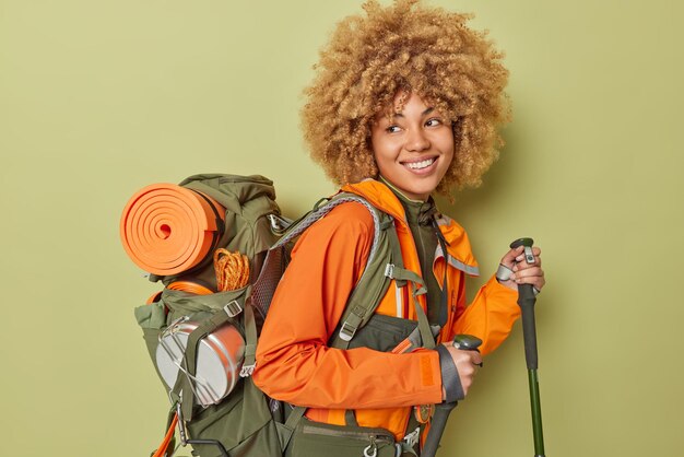 Una excursionista positiva y activa posa con palos escandinavos que intenta llegar a la cima de la montaña vestida con una chaqueta naranja que lleva una mochila y pasa el tiempo libre acampando aislada sobre un fondo verde