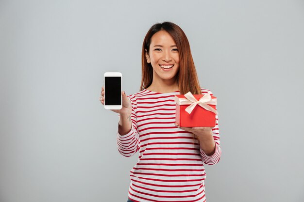 Exhibición asiática feliz de la demostración de la mujer asiática del teléfono que sostiene el regalo.