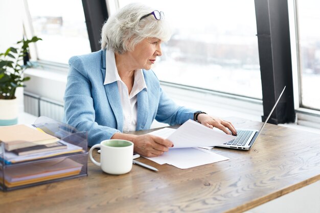 Exitosa abogada anciana con experiencia con un bonito traje y gafas en la cabeza usando una computadora portátil en su lugar de trabajo, mirando la pantalla con una expresión facial concentrada y enfocada