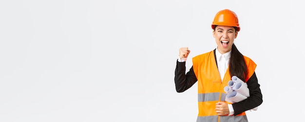 Exitosa arquitecta ingeniera asiática en casco de seguridad y chaqueta reflectante lleva planos del proyecto de construcción y bomba de puño en celebración gritando sí ganando tierno fondo blanco