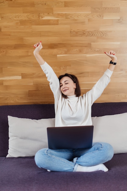 Exitosa dama está trabajando en la computadora portátil sentado en la cama oscura frente a la pared de madera en casa