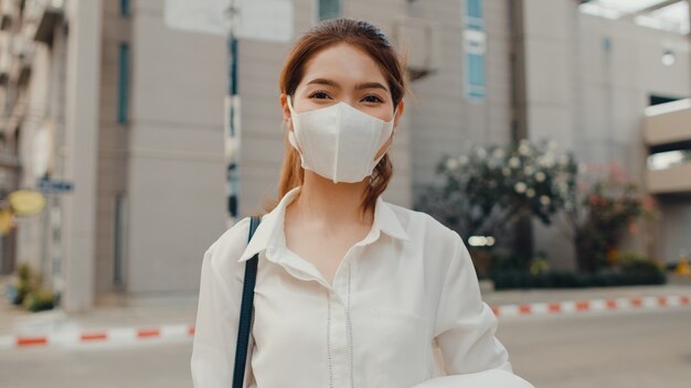 Exitosa empresaria asiática joven en ropa de oficina de moda con mascarilla médica sonriendo en la calle