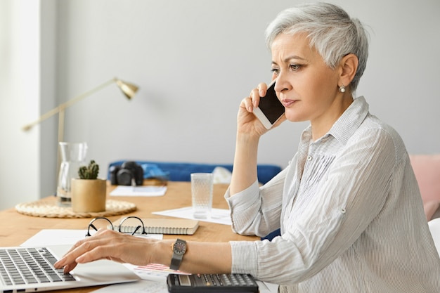 Exitosa empresaria madura confiada con el pelo corto gris que trabaja en el elegante interior de la oficina, usando una computadora portátil y una calculadora, hablando con un socio comercial a través de un teléfono celular. Personas, edad y ocupación