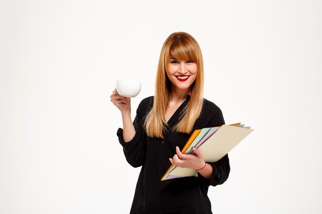 Exitosa empresaria sosteniendo carpetas y taza sonriendo sobre pared blanca