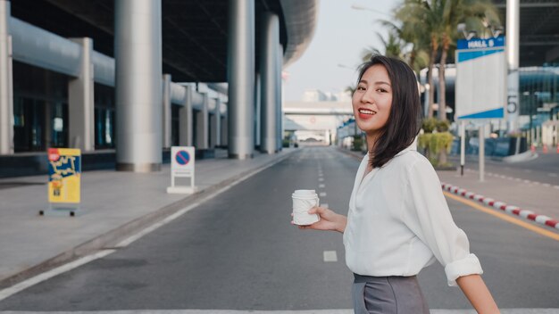 Exitosa joven empresaria de Asia en ropa de oficina de moda sosteniendo un vaso de papel desechable de bebida caliente y usando un teléfono inteligente mientras camina al aire libre en una ciudad moderna urbana