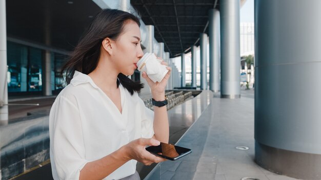 Exitosa joven empresaria de Asia en ropa de oficina de moda sosteniendo un vaso de papel desechable de bebida caliente y usando un teléfono inteligente mientras está de pie al aire libre en una ciudad moderna urbana