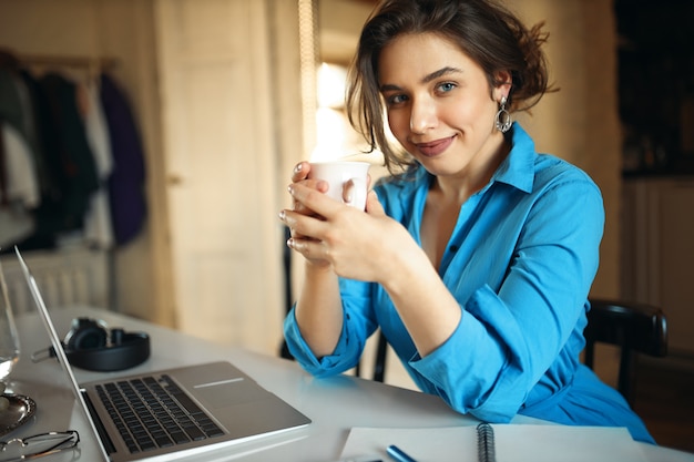 Exitosa maestra joven en vestido de pegamento sentada frente a la computadora portátil, sosteniendo una taza, disfrutando de un café, preparándose para la lección en línea, disfrutando del trabajo a distancia. Chica bonita estudiante con ordenador portátil