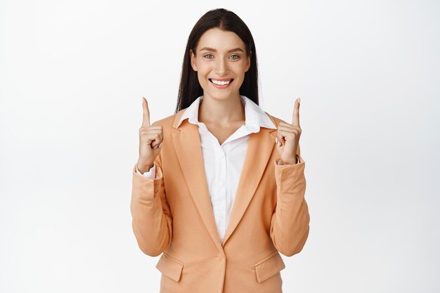 Exitosa mujer corporativa apuntando con el dedo hacia arriba sonriendo y mostrando el nombre de la marca de la empresa de publicidad hacia arriba de pie sobre fondo blanco.