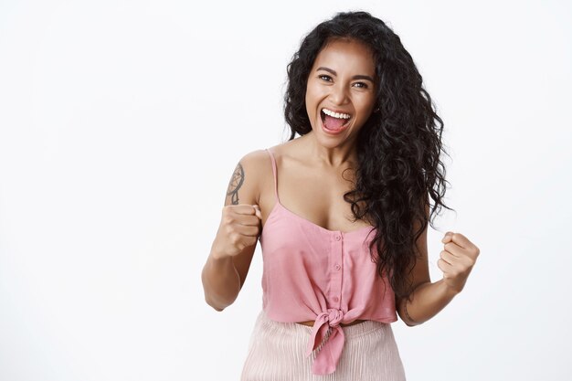 Exitosa mujer joven atractiva, segura de sí misma, con peinado rizado, los tatuajes hacen bombear el puño. gritando, sí, animado, celebrando la victoria, pared blanca