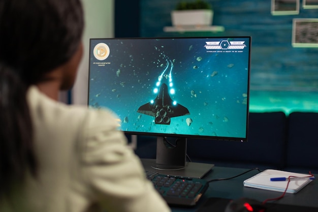 Exitosa mujer jugadora sosteniendo un controlador jugando videojuegos de disparos espaciales durante un torneo en línea en un estudio de juegos en casa. Jugador concentrado que utiliza equipos informáticos RGB profesionales.