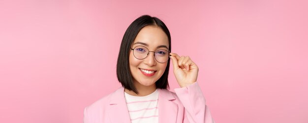 Exitosa mujer de negocios asiática con anteojos sonriendo y mirando profesionalmente a la cámara con traje de pie sobre fondo rosa