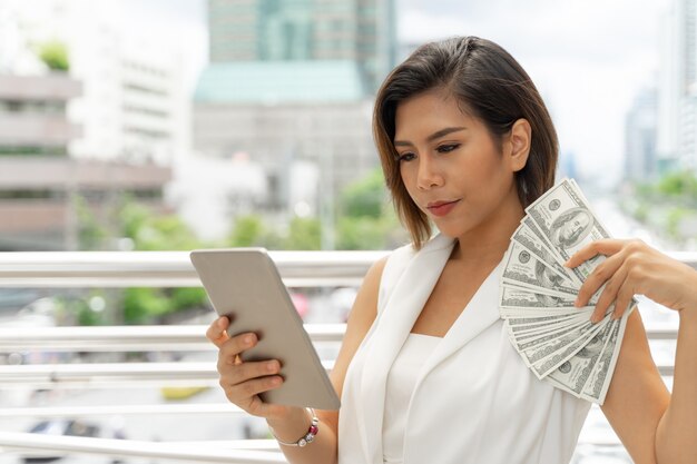 Exitosa mujer de negocios asiática hermosa joven usando teléfono inteligente y dinero billetes de un dólar estadounidense en la mano