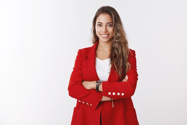 Exitosa mujer de negocios atractiva con chaqueta roja, brazos cruzados, confiada, sonriente, segura de sí misma, asertiva, sabiendo cómo funcionan los clientes, gestionando su propio negocio, pared blanca