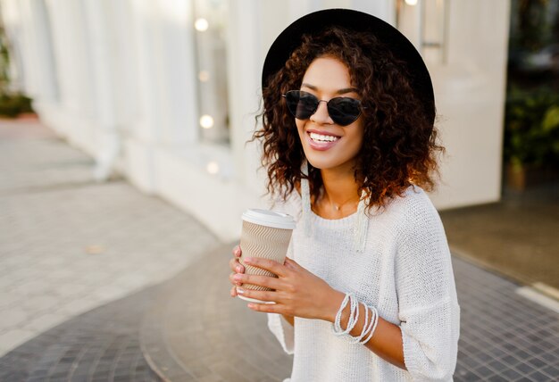 Exitosa mujer negra, blogger o gerente de la tienda hablando por teléfono móvil durante el descanso para tomar café. Sentado en las escaleras y sosteniendo la taza de papel de bebida caliente.