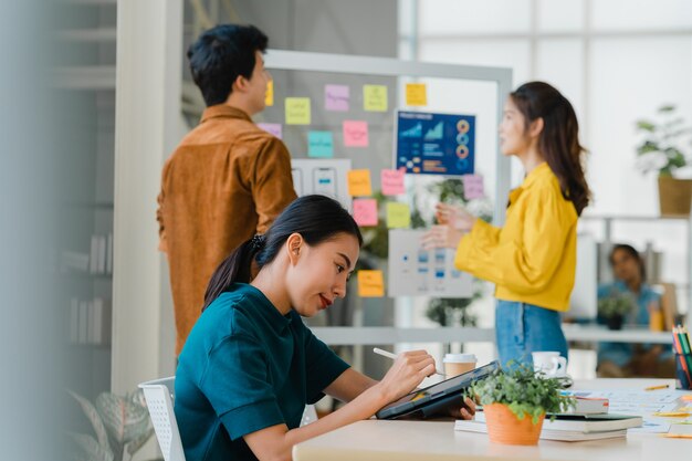 Exitoso ejecutivo Asia joven empresaria elegante ropa casual dibujando, escribiendo y usando lápiz con tableta digital pensando en el proceso de trabajo de ideas de búsqueda de inspiración en la oficina moderna.