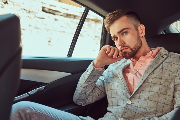 Exitoso y elegante hombre de negocios joven con traje gris y camisa de ping en un asiento trasero en un auto de lujo.