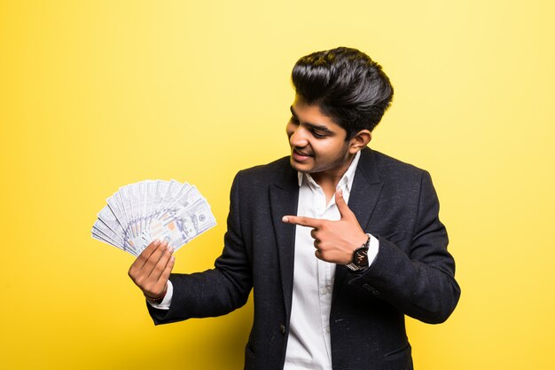 Exitoso empresario indio con billetes de dólar en la mano traje clásico mirando a la cámara con una sonrisa dentuda mientras está de pie contra la pared amarilla