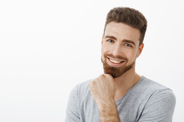 Exitoso y feliz empresario masculino seguro de sí mismo frotando la barba sonriendo y mirando encantado con lindos ojos azules y satisfecho con los buenos resultados de la compañía contra la pared gris