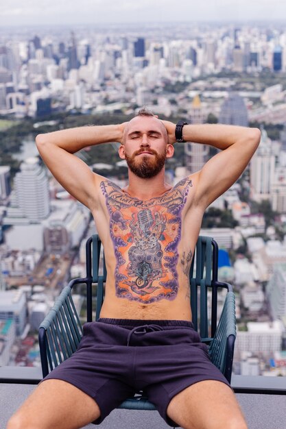 Exitoso guapo europeo barbudo brutal tatuado hombre fuerte en topless con reloj se sienta en una silla en piso alto con increíble vista de la ciudad