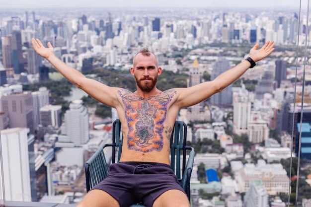 Exitoso guapo europeo barbudo brutal tatuado hombre fuerte en topless con reloj se sienta en una silla en piso alto con increíble vista de la ciudad