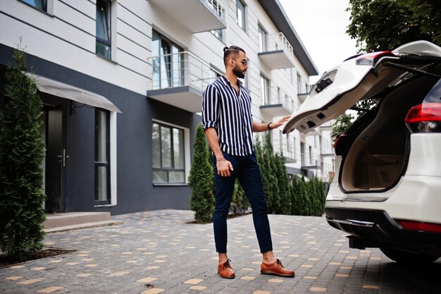 Exitoso hombre árabe vestido con camisa a rayas y gafas de sol baúl cerrado de su automóvil suv blanco Elegantes hombres árabes en transporte