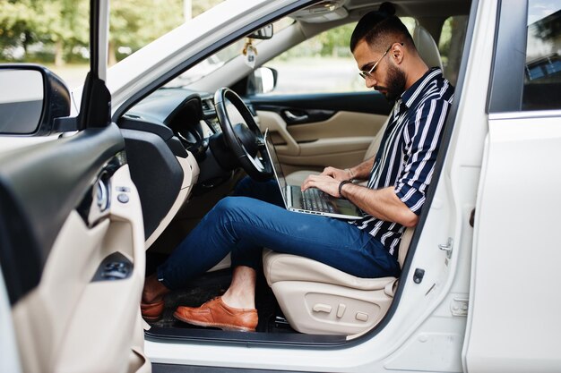 Exitoso hombre árabe vestido con camisa a rayas y gafas de sol posan al volante de su auto todoterreno blanco con una computadora portátil en las manos