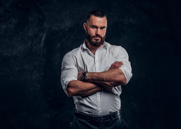 Un exitoso hombre de negocios barbudo con camisa blanca posa en un estudio fotográfico oscuro.