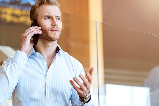 El exitoso hombre de negocios caucásico inteligente que camina en la escalera hace que la comunicación con el teléfono inteligente se comunique con felicidad y un entusiasta fondo de oficina moderno