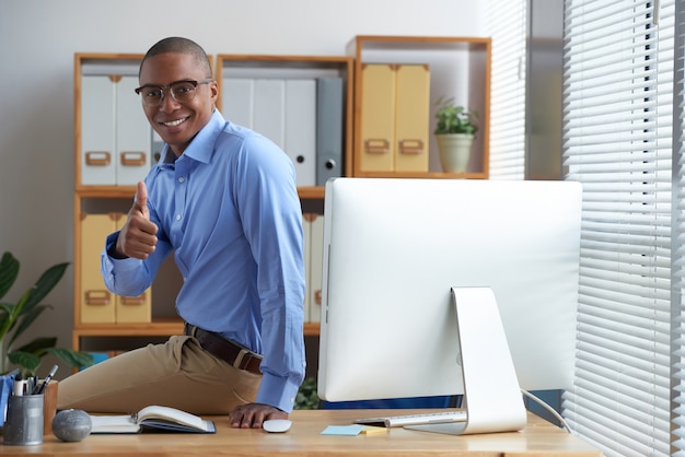 Exitoso hombre de negocios mostrando los pulgares hacia arriba y sonriendo posado en su escritorio de trabajo