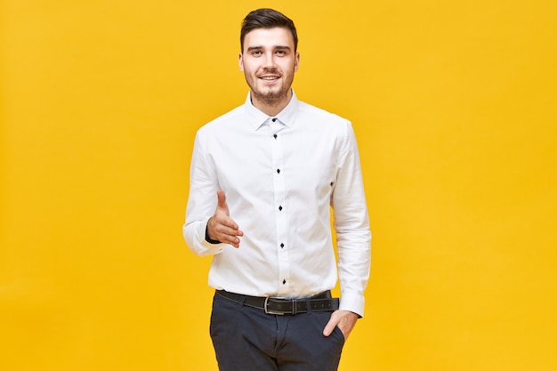 Exitoso joven confiado en camisa blanca formal y pantalón clásico sonriendo y extendiendo la mano para estrechar la suya, haciendo un gesto de bienvenida y saludo, listo para llegar a un acuerdo