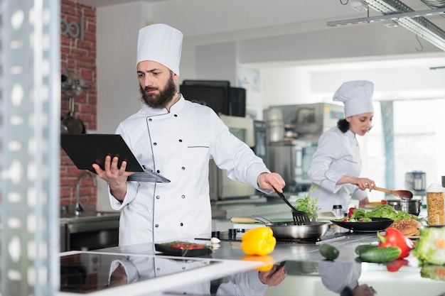 Experto gastronómico preparando guarnición de platos con ingredientes orgánicos frescos mientras sostiene una laptop con una mano. Chef de cocina gourmet cocinando el plato de la cena mientras usa la computadora para seguir la receta de cerca