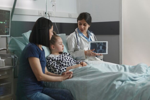 Experto en radiología examinando la imagen de exploración de radiografía de una niña enferma descansando en la sala de pediatría del hospital. Radiólogo que muestra el resultado del escaneo de rayos X de la condición cerebral del niño bajo tratamiento.