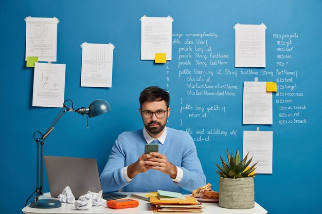 Experto en tecnología digital o entusiasta obsesionado con su trabajo, usa el teléfono móvil, trabaja con dispositivos modernos, rodeado de muchos papeles, posa en el escritorio