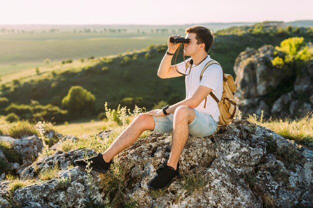 Explorador masculino sentado en la roca mirando a través de binoculares