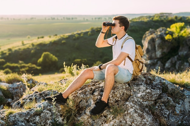 Explorador masculino sentado en la roca mirando a través de binoculares