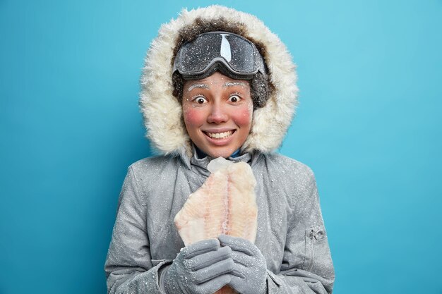 La exploradora polar femenina va a pescar en el hielo durante el invierno vestida con ropa de abrigo sostiene pescado congelado vestido con un atuendo cómodo sobre la pared azul tiembla durante los días fríos preparados para los cambios climáticos